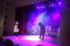 Na scenie dwóch aktorów w przebraniach. Za nimi scenografia przedstawiająca drzewa i polanę z kwiatami. Scenę oświetla fioletowe światło. 