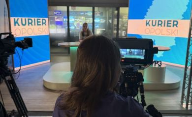 Na zdjęciu młoda dziewczyna w koszulce projektowej Równać Szanse 2025. Stoi za kamerą. W oddali studio nagrań Kuriera Opolskiego. - powiększ