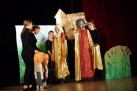 Na zdjęciu widać scenę teatralną, na której kilka osób ubranych na czarno prowadzi duże kukły w barwnych kostiumach przed malowaną scenografią. Przedstawienie ma charakter bajkowy, z elementami fantazji i kolorowymi dekoracjami.