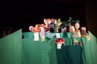 Na zdjęciu widać scenę teatralną z kolorowymi kukiełkami przypominającymi owady w bajkowej scenerii z grzybkami, kwiatami i drzewem. Dekoracje są wykonane z materiałów i papieru, tworząc fantazyjny, dziecięcy klimat.