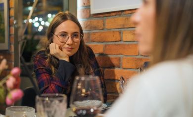 Młoda dziewczyna  w okularach, podpiera brodę prawa ręką, patrzy przed siebie.  Na stole widać talerze, kieliszek z winem i inne naczynia. 