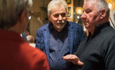 Dwóch mężczyzn ubranych na ciemno rozmawia. Jeden z nich unosi rękę. Przed nimi soi kobieta w okularach, w czerwonym ubraniu. 