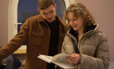 Młody mężczyzna ubrany w brązową kurtkę oraz moda kobieta ubrana w szarą kurtkę przeglądają książkę.