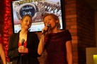 Na zdjęciu dwie wokalistki. Jedna śpiewa do mikrofonu zbliżonego do ust. Druga zaś trzyma mikrofon w dłoniach. Za nimi wyświetlany plakat IV Festiwalu Jazzowego oraz ceglane ściany z czerwonymi światełkami zawieszonymi w rogu tych ścian. 