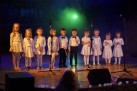 Na zdjęciu widoczna jest grupa dzieci, prawdopodobnie przedszkolaków, stojących na scenie. Wszystkie dzieci ubrane są w białe stroje. Chłopcy mają białe koszule z muszkami, czarne spodnie i trzymają drewniane pałeczki, a dziewczynki mają białe sukienki. Na scenie znajdują się również mikrofony na statywach, co sugeruje, że jest to występ artystyczny.