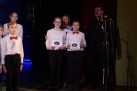 Na zdjęciu najprawdopodobniej uwieczniono ceremonię wręczenia nagród lub dyplomów. W centrum znajdują się dzieci, które otrzymały wyróżnienia, a towarzyszą im dorośli. 