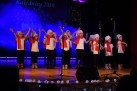 Zdjęcie przedstawia występ grupy dzieci na scenie. W tle widoczny jest baner z napisem „Kolednicy 2019”. Dzieci są ubrane w jednakowe stroje, ciemne spodnie, białe koszulki, czerwone szaliki i białe czapki. 