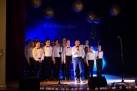 Zdjęcie przedstawia grupę dzieci występujących na scenie podczas wydarzenia 'Kolędnicy 2019'. Dzieci są ubrane w białe koszule, w przypadku chłopców mają dodatkowo czerwone muszki, śpiewają do mikrofonu. Za nimi widoczny jest napis 'Kolędnicy 2019' oraz dekoracje, w tym duże, okrągłe ozdoby wiszące pod sufitem. 