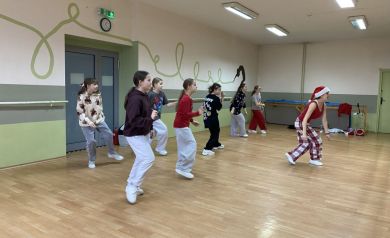 Zdjęcie przedstawia grupę młodych ludzi, dziewcząt, uczestniczących w zajęciach tanecznych w sali tanecznej. Jedna z kobiet ma na głowie świąteczną czapkę. 