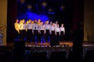 Na zdjęciu widać grupę dzieci i nastolatków, prawdopodobnie chór, występujących na scenie. Dzieci są ubrane w białe koszule i czerwone muchy (chłopcy), stoją na scenie oświetlonej niebieskim światłem. Z tyłu, na wyświetlaczu, widoczny jest napis „Kolędnicy 2019”. Na scenie znajdują się mikrofony, a z przodu widoczne są lampki, które tworzą świąteczny nastrój. 