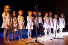 Na zdjęciu grupa dzieci biorąca udział w przeglądzie ,,Kolędnicy 2019'. Dzieci są ubrane w jasne stroje, a dwójka chłopców w czarne spodnie, białe koszule i muszki. Występują na scenie z mikrofonami.