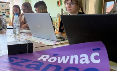 Młodzież siedząca przy stole z laptopami podczas zajęć związanych z projektem. Widać również plakat promujący projekt. - powiększ