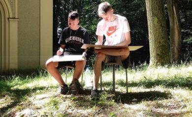 Dwóch młodych mężczyzn siedząc na stołkach, malują obrazy trzymane na kolanach. Znajdują się wśród zieleni. Widać również mury kaplicy. 