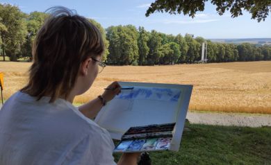 Na pierwszym planie uczestnik wydarzenia rysuje rozpościerający się na drugim planie widok, a składa się na niego zielona aleja drzew, pole pokryte zbożem, ołtarz papieski oraz niebieskie niebo.