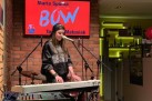 Dziewczyna w długich włosach z czapką na głowie i ciemnej bluzie gra na keyboardzie. Na zdjęciu widać również sprzęt muzyczny, ekran wyświetlający grafikę związaną z odbywającym się wydarzeniem oraz część baru.  