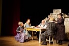 Aktorzy podczas odgrywania sceny teatralnej. Siedząprzy stole. Sceneria sugeruje, że może to być kuchnia. Jedna kobieta stoi. Kobiety ubrane są w długie wzorzyste suknie.