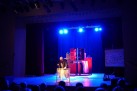 Na zdjęciu aktor podczas odgrywania sceny teatralnej. Są tam również dekoracje potrzebne do spektaklu. Na zdjęciu widać część publiczności. Całość oświetla niebieskie światło.