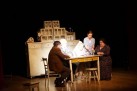 Na zdjęciu scena teatralna. Scenografię stanowi stary regał oraz krzesła i stół, przy którym siedzą aktorzy.