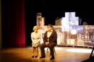 Na zdjęciu scena teatralna. Scenografia ustawiona jest za dwoma aktorami (kobieta i mężczyzna) siedzącymi na krzesłach.