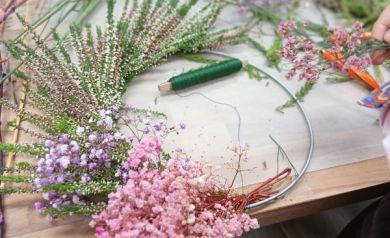 Na zdjęciu widać fragment pracy nad wiankiem florystycznym. Na stole leży metalowa obręcz, do której przytwierdzane są gałązki wrzosu i drobne, kolorowe kwiaty (gipsówka) – w odcieniach różu, fioletu i zieleni. Obok znajduje się zielony drucik florystyczny, używany do mocowania roślin. W tle stoją doniczki z wrzosem i inne przygotowane kwiaty. Po prawej stronie widać rękę osoby, która trzyma małą wiązkę kwiatów gotowych do dołączenia do wianka.