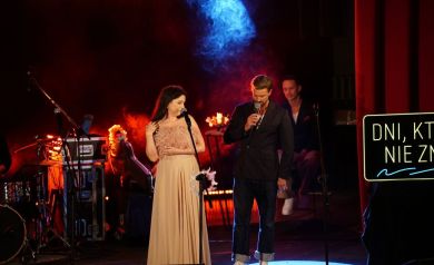 Na zdjęciu widac dwóch mężczyzn i kobietę ubraną w długa elegancką suknię. Jeden z mężczyzn jest na drugim planie. Tło na scenie stanowią instrumenty i sprzęt muzyczny oraz czerwono-niebieskie kolory z ciężkim dymem.