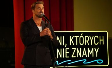Na zdjęciu mężczyzna w czarnej marynarce trzymający statyw z mikrofonem, prawdopodobnie śpiewa. Za nim widnieje podświetlony led.