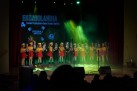 Na zdjęciu grupa przedszkolaków. Stoją w rzędzie. Dziewczęta ubrane są w takie same stroje, chłopcy również. Za nimi osoba dorosła, zaś na wyświetlaczu widnieje plakat związany z odbywającym się wydarzeniem. Widać również część publiczności oraz stojące przed dziećmi na scenie mikrofony. 