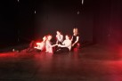 Aktorki podczas sceny teatralnej. Siedzą na podłodze. Scenę oświetla czerwone światło.