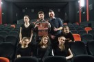 Na zdjęciu grupa młodzieży pozująca do zdjęcia grupowego. Znajdują się na widowni, część siedzi na krzesłach inni stoją. Jeden z chłopaków trzyma w dłoniach plakat i prezentuje go do aparatu. 