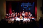 Grupa przedszkolaków na scenie oraz kilku dorosłych. Widać także część publiczności, maskotki i wyświetlany duży napis ,,Ekozoolandia 4. Festiwal Przedszkolaka Gminy Strzelce Opolskie POZNAJ MISIA'.  