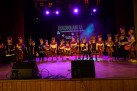 Zdjęcie przedstawia grupę przedszkolaków stojących na scenie. Mają przebrania i maski na głowach. W tle wyświetlany jest plakat odbywającej się imprezy. Scenę oświetla fioletowe światło. 