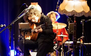 Kobieta grająca na gitarze. Przed nią ustawiony został mikrofon. Obok widać lampę stojącą z abażurem. W tle widać perkusistkę grająca na zestawie perkusyjnym.