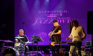 Na zdjęciu trzej mężczyźni grający na instrumentach. W tle wyświetlany jest plakat dotyczący XI Festiwalu - Strzeleckie Ogrody Jazzowe. 