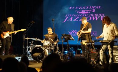 Zdjęcie przedstawia koncert muzyczny podczas XI Festiwalu ,,Strzeleckie Ogrody Jazzowe'. Na scenie widoczny jest czteroosobowy zespół muzyczny podczas występu. Tło sceny jest niebieskie, a na nim widnieje wyświetlany napis 'XI FESTIWAL STRZELECKIE OGRODY JAZZOWE'. W skład zespołu wchodzą: gitarzysta elektryczny (po lewej), perkusista (w centrum, za zestawem perkusyjnym), basista (po prawej od perkusisty), wibrafonista. W dolnej części kadru widać sylwetki publiczności. Scena jest profesjonalnie oświetlona, a muzycy korzystają ze standardowego sprzętu muzycznego, w tym mikrofonów i nagłośnienia.