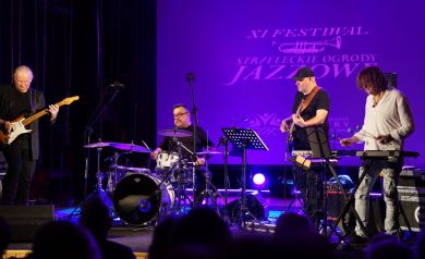 Zdjęcie przedstawia koncert muzyczny podczas XI Festiwalu ,,Strzeleckie Ogrody Jazzowe'. Na scenie widoczny jest czteroosobowy zespół muzyczny podczas występu. Tło sceny jest fioletowe, a na nim widnieje wyświetlany napis 'XI FESTIWAL STRZELECKIE OGRODY JAZZOWE'. W skład zespołu wchodzą: gitarzysta elektryczny (po lewej), perkusista (w centrum, za zestawem perkusyjnym), basista (po prawej od perkusisty), wibrafonista. W dolnej części kadru widać sylwetki publiczności. Scena jest profesjonalnie oświetlona, a muzycy korzystają ze standardowego sprzętu muzycznego, w tym mikrofonów i nagłośnienia.
