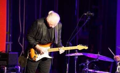 Mężczyzna grający na gitarze elektrycznej. Obok niego stoi sprzęt muzyczny, taki jak perkusja, czy mikrofon. 