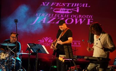 Zdjęcie przedstawia zespół muzyczny występujący na żywo podczas XI Festiwalu Strzeleckie Ogrody Jazzowe. Scena jest oświetlona głównie czerwonym i niebieskim światłem, z widocznym dymem scenicznym. W tle znajduje się wyświetlany plakat z napisem 'XI FESTIWAL STRZELECKIE OGRODY JAZZOWE'. Na scenie widocznych jest trzech muzyków. Po lewej stronie jest perkusista siedzący za zestawem perkusyjnym. W centrum jest basista z czapką bejsbolową, grający na gitarze basowej. A także Bernard Maseli.


