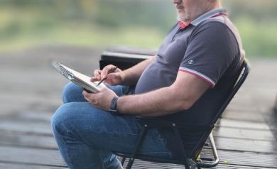 Mężczyzna w jeansach, w ciemnej bluzce polo, z kaszkietem na głowie siedzi na krześle i szkicuje. W tle widać zieleń i niebo podczas zachodu słońca.