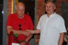 Na zdjęciu dwóch mężczyzn jeden w białej koszuli w wzorki, drugi w czerwonej koszulce, podają sobie prawe dłonie. Jeden z nich (ubrany w czerwoną koszulkę) trzyma czerwoną różę w ręku. 