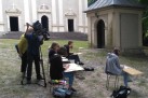 Na zdjęciu widać grupę młodych osób rysujących na świeżym powietrzu przed kapliczką i kościołem, podczas gdy obok nich ekipa filmowa nagrywała materiał. 