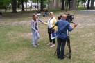 Na zdjęciu kobieta przeprowadzająca wywiad z młodą dziewczyną, mężczyzna kameruje wywiad. W oddali widać drzewa i innych uczestników wydarzenia. 