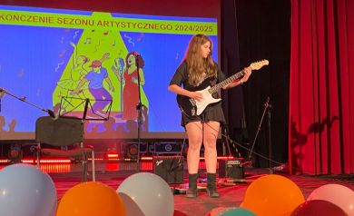 Dziewczyna grająca na gitarze elektrycznej. Wokół niej sprzęt muzyczny: wzmacniacze, mikrofony. Przed gitarzystką widać kolorowe balony. Zaś za nią wyświetlana jest grafika związana z Zakończeniem Sezonu Artystycznego 2024/2025. 