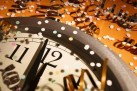 Na zdjęciu widać fragment dużego zegara, którego wskazówki pokazują godzinę tuż przed północą lub przed południem. Zegar ma srebrną ramę i klasyczną tarczę z czarnymi cyframi. Wokół zegara rozsypane są kolorowe konfetti i złote serpentyny, które nadają obrazowi sylwestrowo-karnawałowy charakter. Tło jest w ciepłym, pomarańczowym odcieniu.