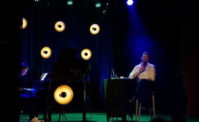 Dwóch muzyków na scenie, jeden śpiewa drugi gra na fortepianie. Lampy rzucające światło w rożnych kolorach na artystów. 