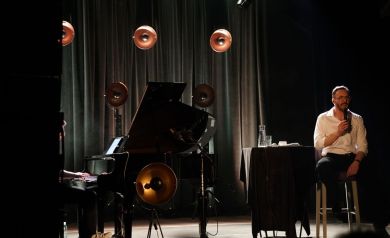 Muzycy na scenie (piosenkarz i pianista). Na drugim planie widnieją lampy do oświetlania sceny i artystów. 