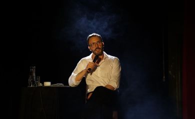 Sławek Uniatowski w białej koszuli z mikrofonem w ręce na czarnym tle. Opiera się jedną ręką o stolik. 