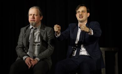 Dwóch mężczyzn w garniturach siedzi koło siebie. Jeden z nich trzyma ręce w pozycji jak by kierował kierownicą. 