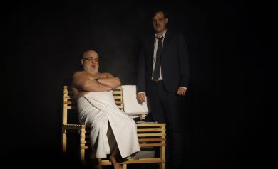 Mężczyzna owinięty ręcznikiem siedzi na drewnianej ławie. Obok niego stoi mężczyzna w garniturze.
