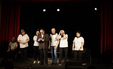 Siedmioro aktorów podczas swojego występu. Prawie wszyscy ubrani są w białe koszulki, jedynie jeden z nich ma na sobie marynarkę i spodnie garniturowe.