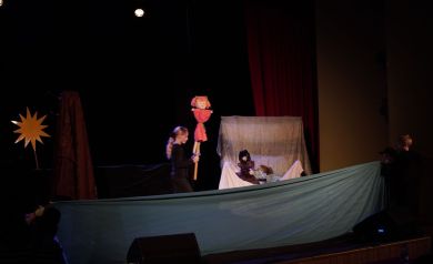 Na pierwszym planie widoczna jest dekoracja ustawiona na scenie. Jest to m.in. niebieski materiał rozciągnięty na prawie całą długość sceny. Poza tym można dostrzec kilku aktorów w tym ubraną na czarno dziewczynkę, która niesie kukłę.